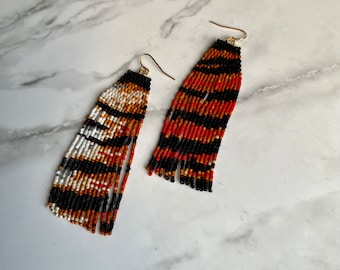 May include: A pair of long, beaded earrings with a geometric pattern in black, white, orange, and brown. The earrings are made of small, round beads and have a tassel-like fringe at the bottom.