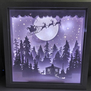 May include: A white papercraft shadow box with a purple background depicting a winter scene. Santa Claus is flying in his sleigh pulled by reindeer through a snowy forest with a cabin in the distance. The moon is full and there are stars in the sky.