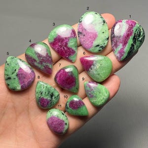 Puede incluir: Una colección de once piedras preciosas de zoisita pulidas con inclusiones de rubí. Las piedras varían en forma, incluyendo ovaladas, de lágrima e irregulares. Las piedras preciosas muestran una mezcla de colores verde y magenta, con algunas inclusiones negras.