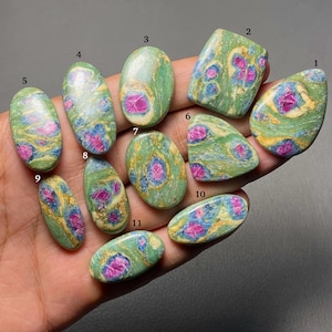 May include: Eleven polished cabochon gemstones labeled one through eleven, featuring a green base with vibrant pink and blue inclusions. Each stone displays a unique marbled pattern with swirling colors, presented in various shapes including oval, pear, and rectangular.