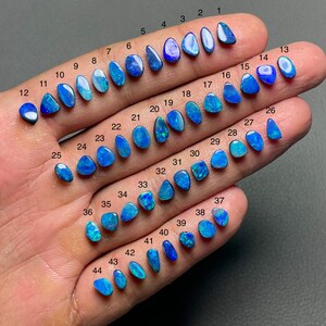 May include: A collection of 44 blue opal gemstones, each with a unique shape and pattern. The opals are numbered and displayed on a hand, showcasing their vibrant colors and variations. The gemstones range in size and shape, from small teardrops to larger, irregular forms.