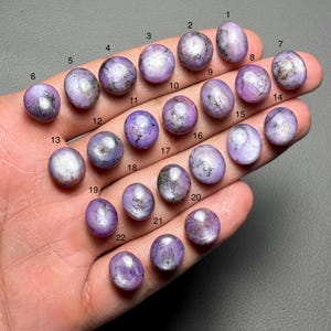 May include: A collection of twenty-two oval-shaped, polished gemstones in varying shades of lavender and purple. Each stone has unique patterns and inclusions, with a glossy finish. The gemstones are numbered 1 through 22.