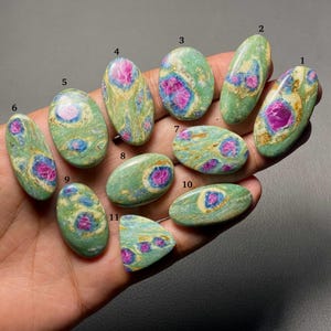 May include: Eleven polished oval and triangular cabochons featuring a green fuchsite matrix with embedded magenta ruby crystals. Each stone is numbered from 1 to 11, displaying unique patterns of green, gold, and vibrant pink mineral inclusions.