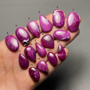 Könnte beinhalten: Eine Sammlung von fünfzehn polierten, ovalen und tropfenförmigen Edelsteinen in verschiedenen Größen. Die Steine sind tiefmagenta mit weißen und schwarzen Adern. Die Edelsteine werden auf einem neutralen Hintergrund präsentiert.