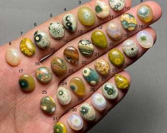 Könnte beinhalten: Eine Sammlung von einunddreißig polierten Edelsteinen in verschiedenen Formen und Farben, darunter Grün, Gelb, Braun und Weiß. Jeder Stein hat einzigartige Muster und Texturen, die auf einer Hand präsentiert werden.