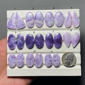 May include: A collection of eleven polished gemstones in various shapes, including triangles, ovals, and teardrops. The stones are primarily shades of purple, with some showing lighter marbling. A US nickel coin is included for size reference.