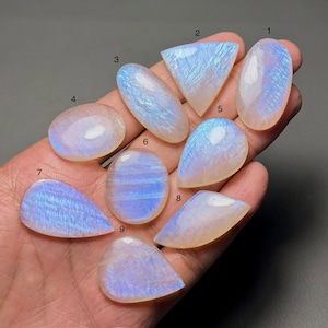 Op de afbeelding: Een verzameling van negen gepolijste maansteen edelstenen in verschillende vormen, waaronder ovaal, druppelvormig en driehoekig. De stenen vertonen een glinsterend, iriserend kleurenspel, voornamelijk in tinten blauw en wit, met een parelmoerglans. De edelstenen worden in een hand gehouden.