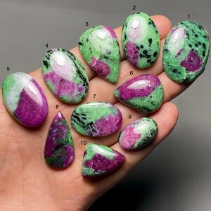 Puede incluir: Una colección de piedras preciosas pulidas de zoisita y rubí en varias formas, incluyendo ovaladas, de lágrima y triangulares. Las piedras presentan una mezcla de tonos verdes y rosas con motas negras, mostrando patrones y texturas naturales. Las piedras preciosas se sostienen en una mano.