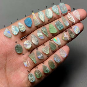 Op de afbeelding: Een verzameling van 34 stukken zeeglas in verschillende vormen en kleuren, waaronder blauw, groen en iriserend. De stukken worden in een hand gehouden en tonen hun unieke texturen en natuurlijke schoonheid. Elk stuk is genummerd.