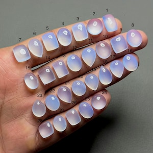 May include: A collection of twenty-eight polished, translucent gemstones in various shapes, including ovals, teardrops, and rectangles. The stones have a soft, milky blue and lavender color, with a subtle sheen. The gemstones are arranged on a neutral background.