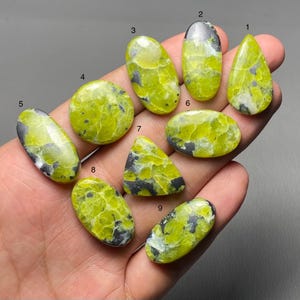 Pode incluir: Uma coleção de nove pedras preciosas polidas em várias formas, incluindo ovais e em forma de gota. As pedras apresentam uma cor amarelo-esverdeada vibrante com inclusões cinza escuro e pretas, criando um padrão único. As pedras preciosas são seguradas em uma mão.