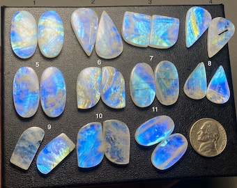May include: A collection of iridescent moonstone cabochons in various shapes, including oval, teardrop, and irregular forms. The stones display a play of blue, white, and yellow colors. A U.S. nickel is included for size reference.