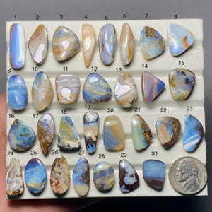 May include: A collection of 30 polished opal gemstones in various shapes and colors, including blue, brown, and white. The gemstones are displayed on a white surface, with a US dime for size comparison.