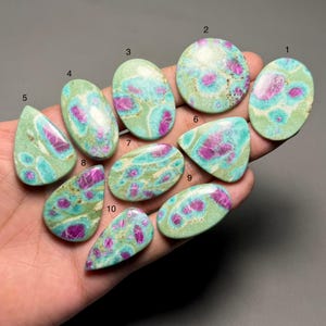 Puede incluir: Una colección de diez piedras preciosas pulidas de diversas formas, incluyendo ovaladas, en forma de lágrima y triangulares. Las piedras presentan un patrón moteado de tonos verdes, turquesa y magenta. Las piedras se muestran sobre un fondo neutro.