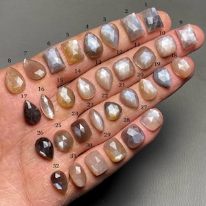 Op de afbeelding: Tweeëndertig regenboogkleurige maansteen-cabochons in verschillende vormen en maten, waaronder druppel-, ovaal- en rechthoekige vormen, gerangschikt in rijen op een hand.