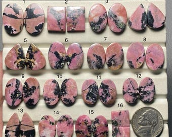 May include: A collection of rhodochrosite gemstones in various shapes, including oval, square, and teardrop. The stones display a mix of pink, black, and white patterns. A US nickel is included for size reference. The gemstones are arranged on a white display.