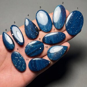 Op de afbeelding: Een verzameling van elf gepolijste blauwe edelstenen met witte spikkels, elk met een unieke vorm. De edelstenen worden in een hand gehouden, waarbij hun verschillende maten en ontwerpen worden getoond. De stenen hebben een glanzende afwerking.