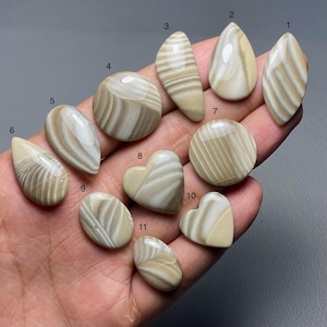 May include: A collection of eleven polished stones in various shapes, including teardrop, oval, and heart. The stones feature a creamy beige and white striped pattern. The stones are displayed on a neutral gray background.