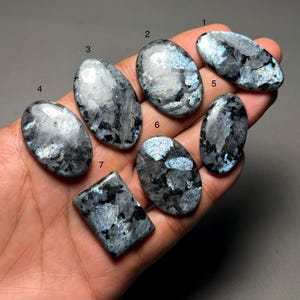 Op de afbeelding: Zeven gepolijste labradoriet edelstenen in verschillende ovale en rechthoekige vormen. De stenen vertonen een mix van grijze, zwarte en iriserende blauwe tinten. De edelstenen worden in een hand gehouden, wat hun natuurlijke schoonheid en unieke patronen laat zien.