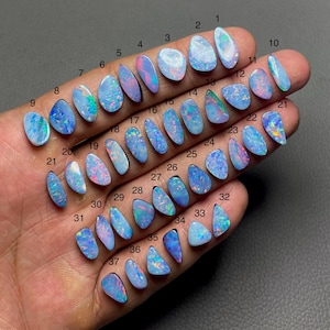 Puede incluir: Una colección de 37 piedras preciosas de ópalo pulidas de diversas formas y tamaños, presentadas en una mano. Los ópalos exhiben una gama de colores, incluyendo azul, rosa y verde, con patrones internos visibles. Cada piedra está numerada.