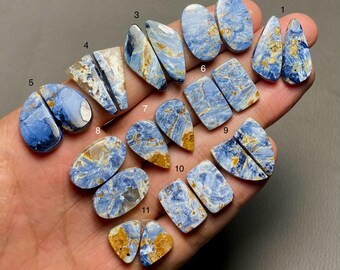 May include: A collection of 11 pairs of blue and gold tiger's eye cabochons, each pair is numbered 1 through 11. The cabochons are various shapes and sizes, including oval, teardrop, and rectangular.