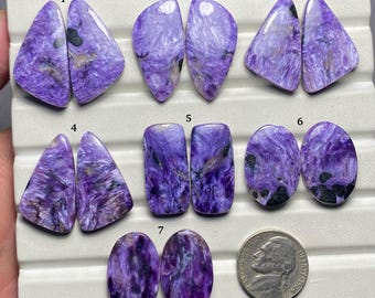 May include: Seven pairs of purple and white spectrolite cabochons, ranging in size and shape. The cabochons are arranged in rows on a white surface, with numbers 1 through 7 labeling each pair.
