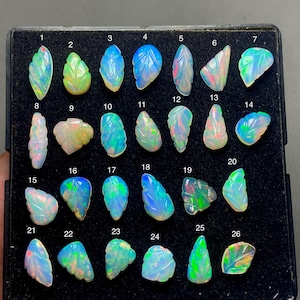 May include: A collection of 26 carved opal gemstones, each with unique leaf-like designs and iridescent colors. The opals are displayed in a black tray, showcasing their individual shapes and vibrant hues of blue, green, and white.