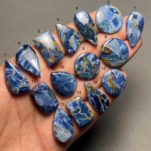 May include: Fifteen polished Pietersite gemstones in diverse shapes, including teardrops, ovals, and rectangles. The stones exhibit a striking blue and gold colour palette, with swirling patterns and natural veining. The gemstones are held in a hand.