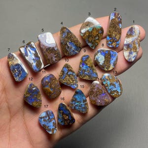 Puede incluir: Colección de diecisiete piedras preciosas de ópalo pulidas, cada una con patrones y colores únicos. Los ópalos muestran una mezcla de tonos azules, dorados y marrones. Las piedras son de forma irregular.