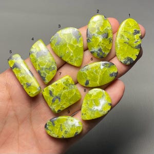 May include: A collection of nine polished gemstones in various shapes, including oval, rectangular, and teardrop. The stones are primarily a vibrant yellow-green color, with gray and black veining patterns throughout. The gemstones are displayed against a neutral background.