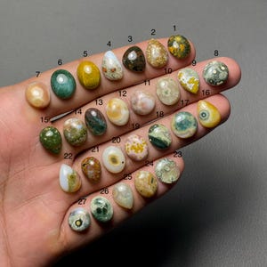 Op de afbeelding: Een verzameling van 27 gepolijste edelstenen in verschillende vormen en kleuren, waaronder ovaal, druppelvormig en rond. De edelstenen worden in een hand gehouden, waarbij hun unieke patronen en texturen worden getoond. Kleuren variëren van groen en geel tot bruin en wit.