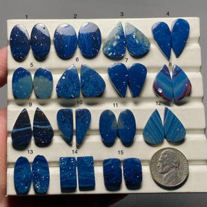 May include: A collection of fifteen pairs of polished blue gemstones in various shapes, including ovals, teardrops, and rectangles. Some stones have white speckles, while others feature swirling patterns. A US nickel is included for size reference.