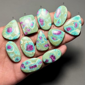 Puede incluir: Una colección de once piedras preciosas pulidas de diversas formas, incluyendo óvalos y rectángulos. Las piedras presentan un color base verde claro con patrones magenta y turquesa. Las piedras preciosas se sostienen en una mano. Medidas en centímetros.