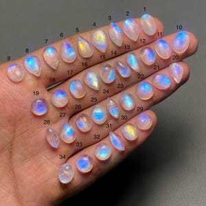 Könnte beinhalten: Eine Hand zeigt 34 polierte Mondstein-Edelsteine in verschiedenen Formen und Größen. Die Steine weisen eine Reihe von Farben auf, darunter Weiß, Blau und Gelb, mit einem schimmernden, irisierenden Effekt. Jeder Stein ist zur Identifizierung nummeriert.