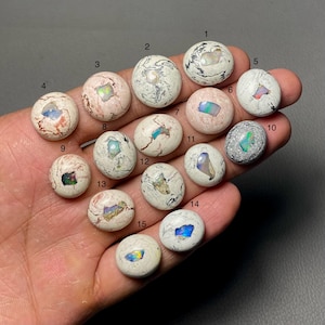 May include: Fifteen polished opal cabochons in various colors and patterns. The opals are set against a neutral background. Each stone is numbered from 1 to 15. The opals have a mix of white, pink, and gray tones with flashes of color.