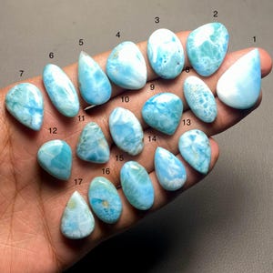 Puede incluir: Una colección de diecisiete piedras preciosas de larimar pulidas en varias formas, incluyendo ovaladas y de lágrima. Las piedras muestran una gama de tonos azul claro con vetas blancas, sostenidas en una mano.