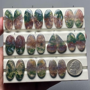 May include: An assortment of eleven moss agate gemstones in various oval and triangular shapes. The stones display a mix of green, brown, and white colors. A US nickel is included for size comparison.