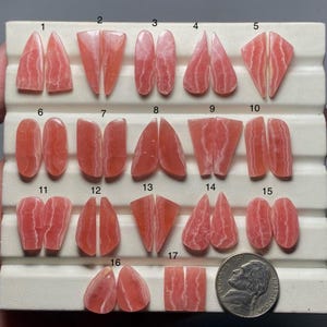 May include: A collection of seventeen pairs of polished, pink rhodonite gemstones, each with unique patterns and shapes. The gemstones are displayed on a white tray, with a US nickel coin for size comparison. The stones vary in size and cut.