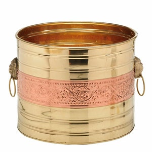 May include: A round, gold-toned metal planter with a copper band featuring an intricate floral design. The planter has two lion head handles with rings.