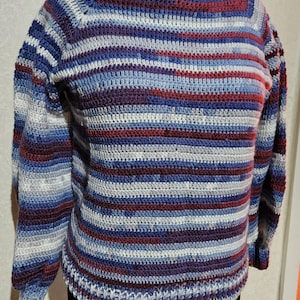 Può includere: Un maglione a righe lavorato a uncinetto in tonalità di blu, viola e rosso. Il maglione ha un collo rotondo e maniche lunghe.