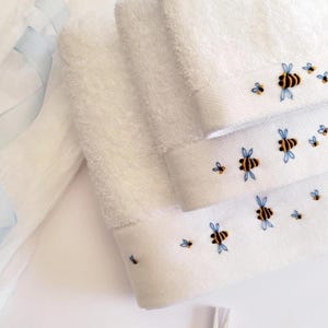 May include: White hand towels with embroidered bee designs. The towels have a soft, textured appearance and feature a border with detailed embroidery of bees in black, yellow, and blue. A light blue ribbon is visible in the background.
