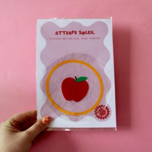 May include: A package featuring a window sticker with an apple design. The sticker has a red apple with a green leaf inside a yellow circle, with the text "ATTRAPE SOLEIL" and "- STICKER ARC-EN-CIEL POUR FENETRE -" in pink and white.