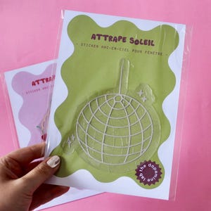 Pode incluir: Um autocolante verde com o texto "ATTRAPE SOLEIL" e "STICKER ARC-EN-CIEL POUR FENETRE" apresenta um design de bola de discoteca. O autocolante está numa manga de plástico transparente. Um segundo autocolante com o texto "ATTRA" é visível no fundo.