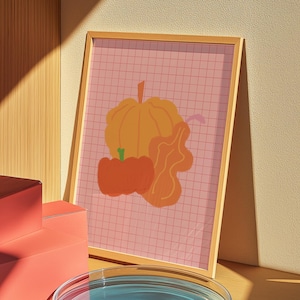 Peut inclure: Une affiche encadrée représentant une illustration stylisée de citrouilles et de courges dans des tons orange et jaune sur un fond rose quadrillé. L'œuvre est présentée dans un cadre en bois, appuyée contre un mur.