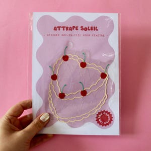 May include: A clear plastic package containing a suncatcher sticker. The sticker is a light beige outline of a heart shape, adorned with red cherry illustrations. The text "ATTAPE SOLEIL - STICKER ARC-EN-CIEL POUR FENÊTRE" is printed at the top.