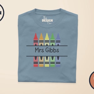 Pode incluir: Uma t-shirt de gola redonda em azul poeirento com um gráfico de cinco lápis de cera coloridos em vermelho, laranja, amarelo, verde e roxo. Os lápis são divididos por uma linha horizontal com o texto preto Mrs Gibbs numa fonte sem serifa.