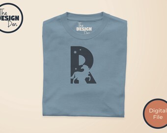 Puede incluir: Una camiseta de color azul polvoriento con un gráfico de la letra R en gris oscuro que contiene una silueta de unicornio blanca y estrellas pequeñas. La etiqueta interior del cuello muestra el texto The Design Den y el logo arriba.