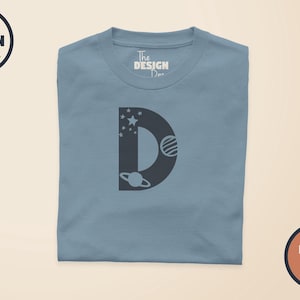 May include: A dusty blue t-shirt featuring a large, dark grey letter D on the front. The letter is decorated with small stars and two planets with rings. The inner neck label displays the text The Design Den, and a logo is in the top left corner.