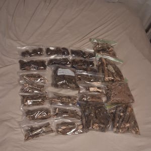 May include: A collection of numerous small, clear plastic resealable bags filled with various pieces of brown, textured tree bark, twigs, and small wooden fragments, arranged in a grid-like pattern on a light-colored fabric surface.