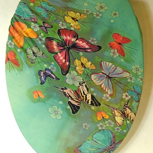 Puede incluir: Asiento de inodoro decorativo con diseño de mariposas. El asiento tiene un fondo turquesa con mariposas coloridas en tonos rojos, naranjas, amarillos y azules. Flores blancas y enredaderas verdes completan el diseño, creando un tema natural.